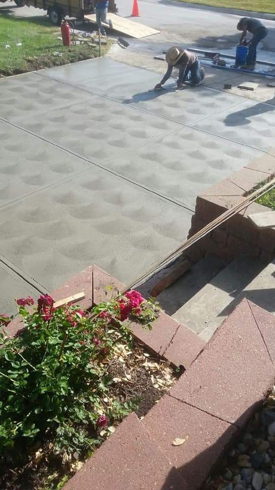 Workers finishing a concrete driveway. Freshly poured cement with two workers, bricks in foreground, and greenery.