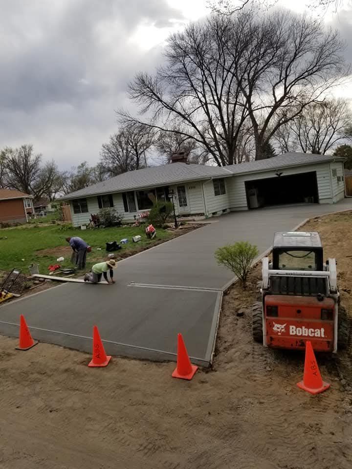 New concrete driveway being installed at a house; workers, orange cones, and Bobcat equipment are visible.