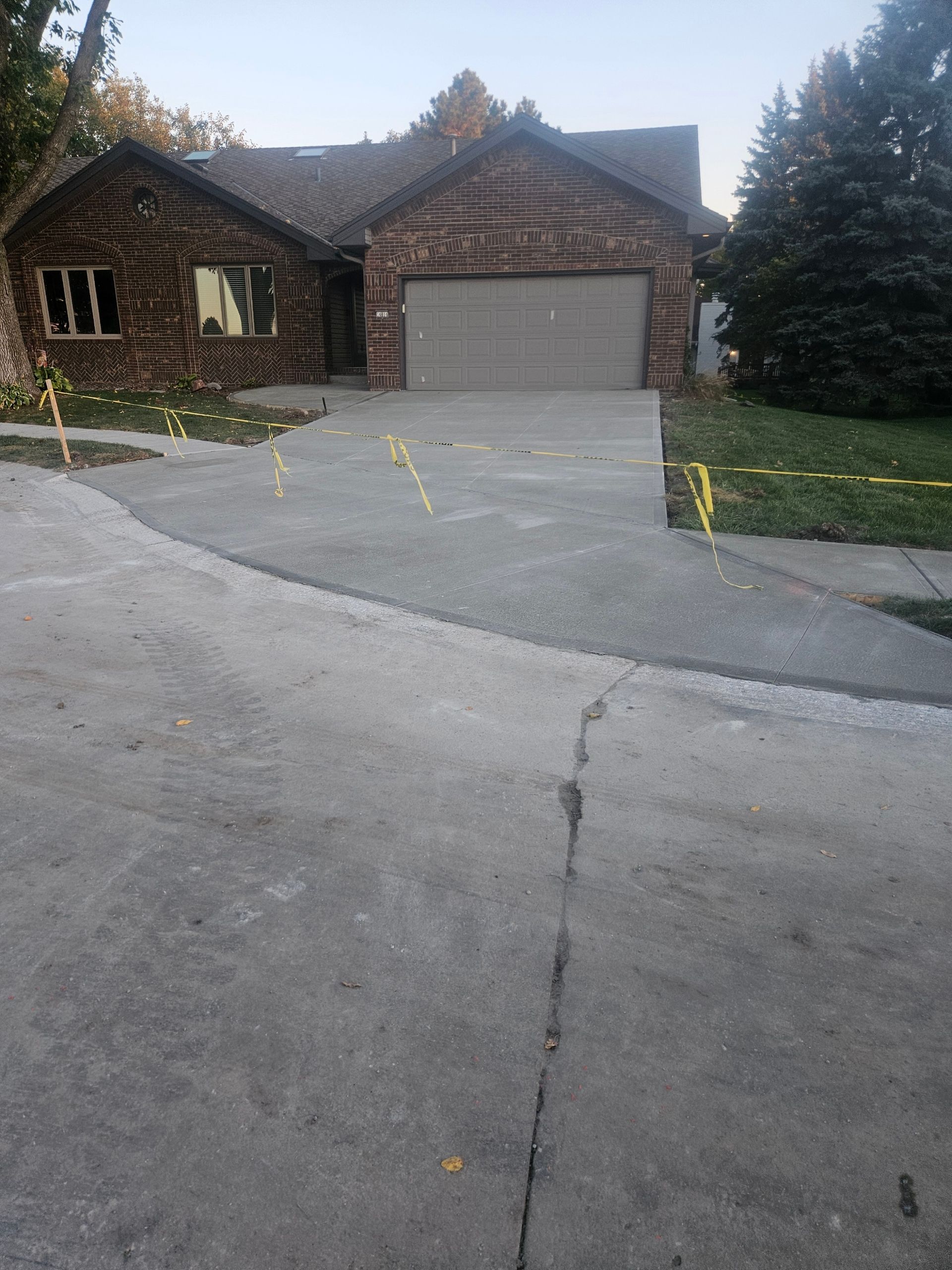 Brick house with a two-car garage and concrete driveway. Asphalt street in foreground with cracks.
