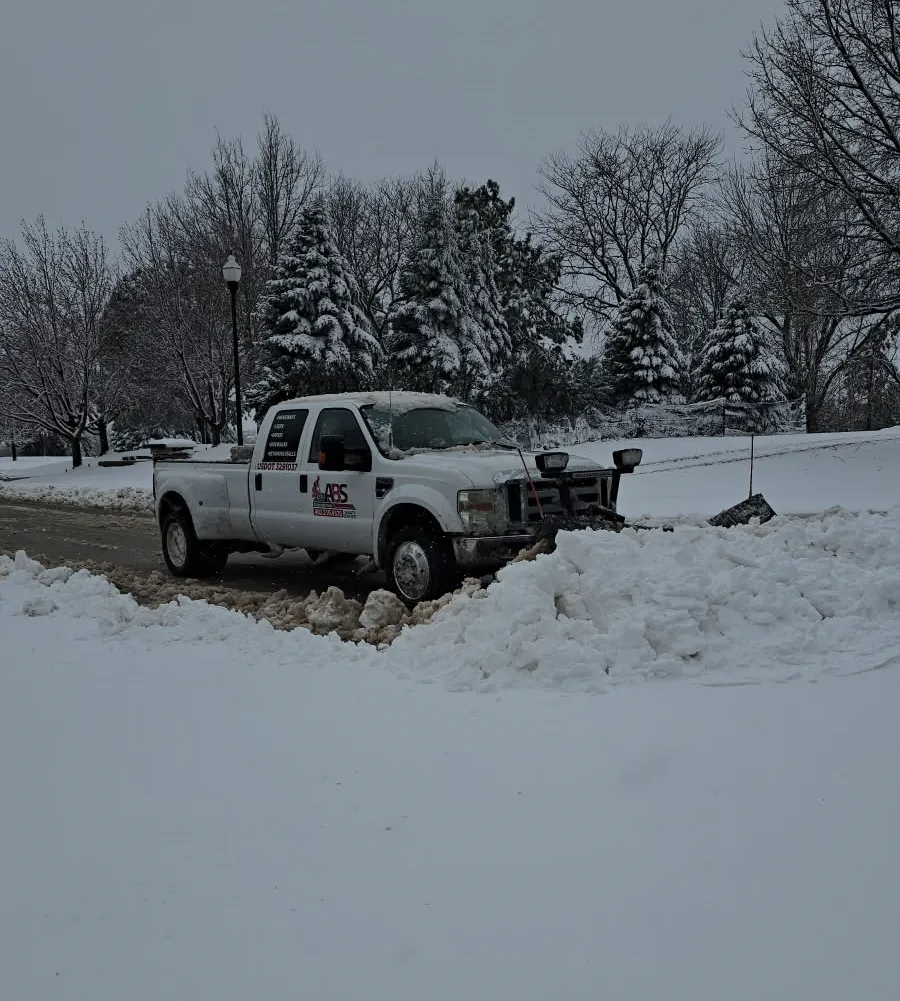 White pickup truck with plow clearing snow from a road on a snowy day.