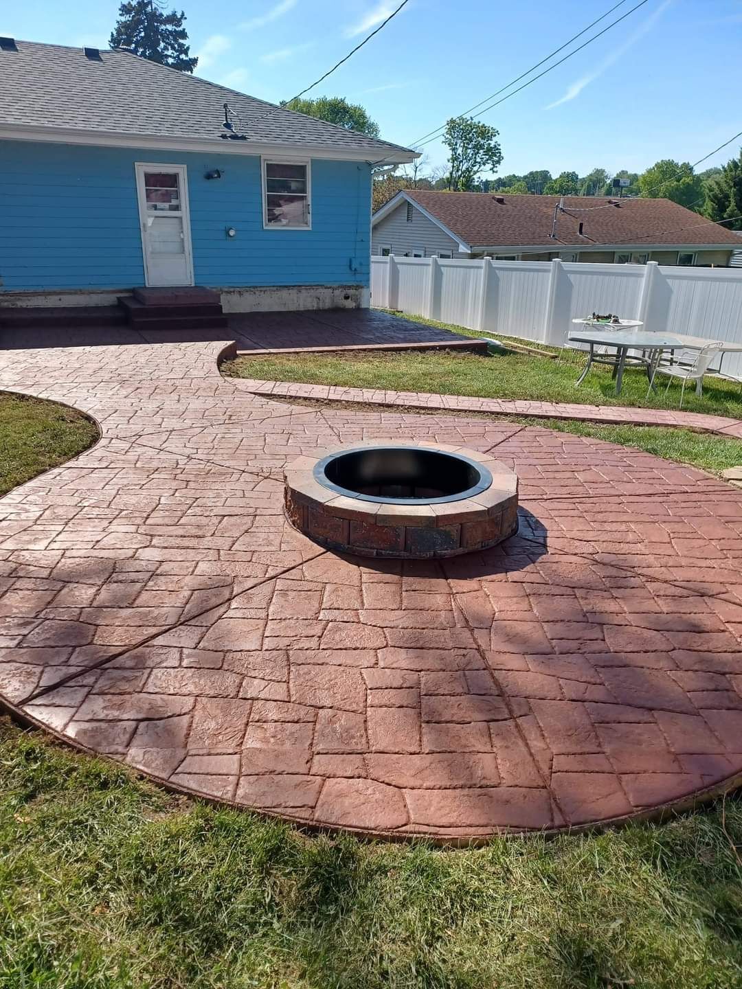 Brown stamped concrete patio with fire pit, blue house, and white fence.