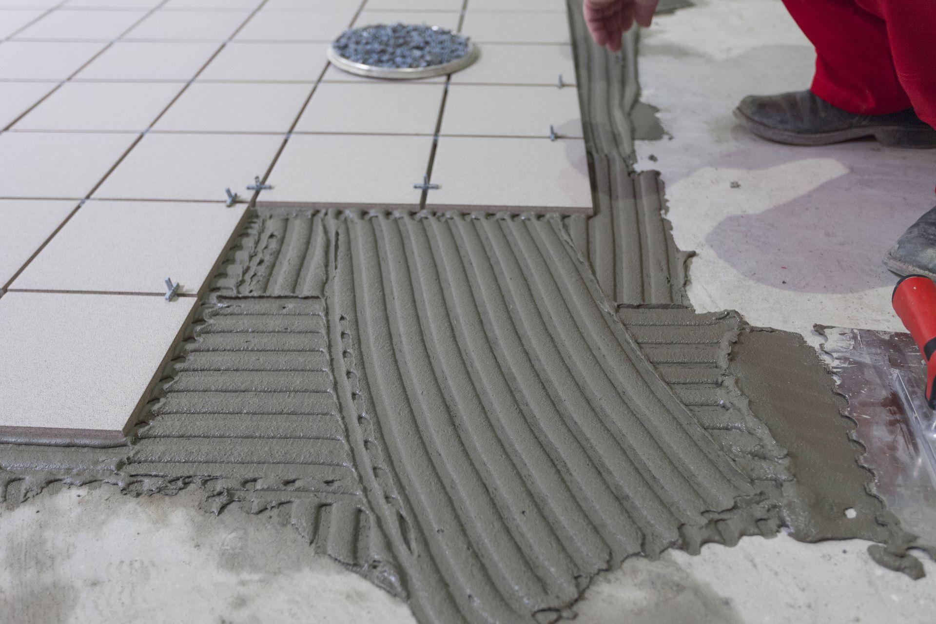 Person installing floor tiles, spreading mortar with notched trowel.  Tiled floor with spacers visible.