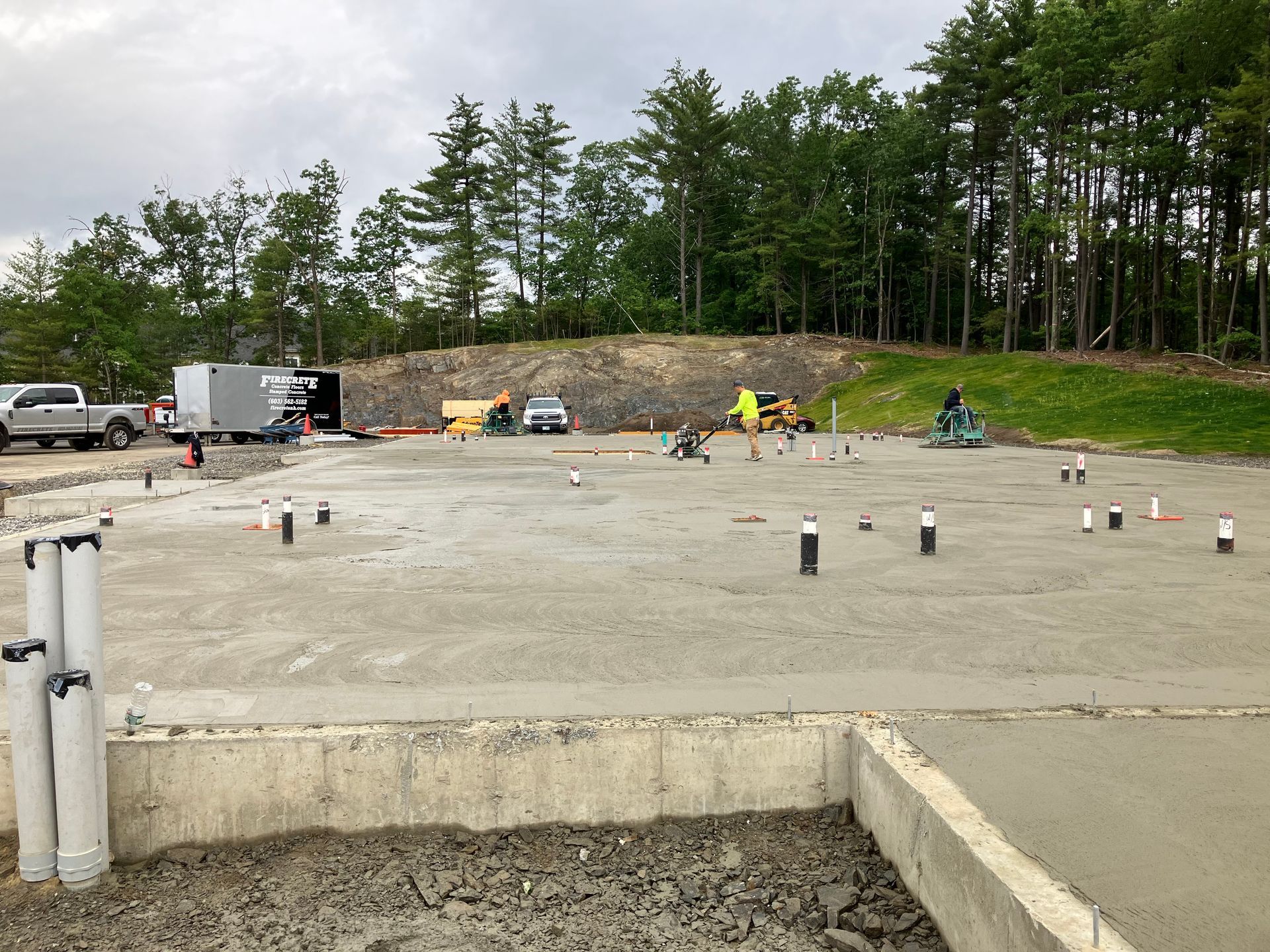 Construction site with concrete foundation and pipes, workers, equipment, and a tree-lined hillside in the background.