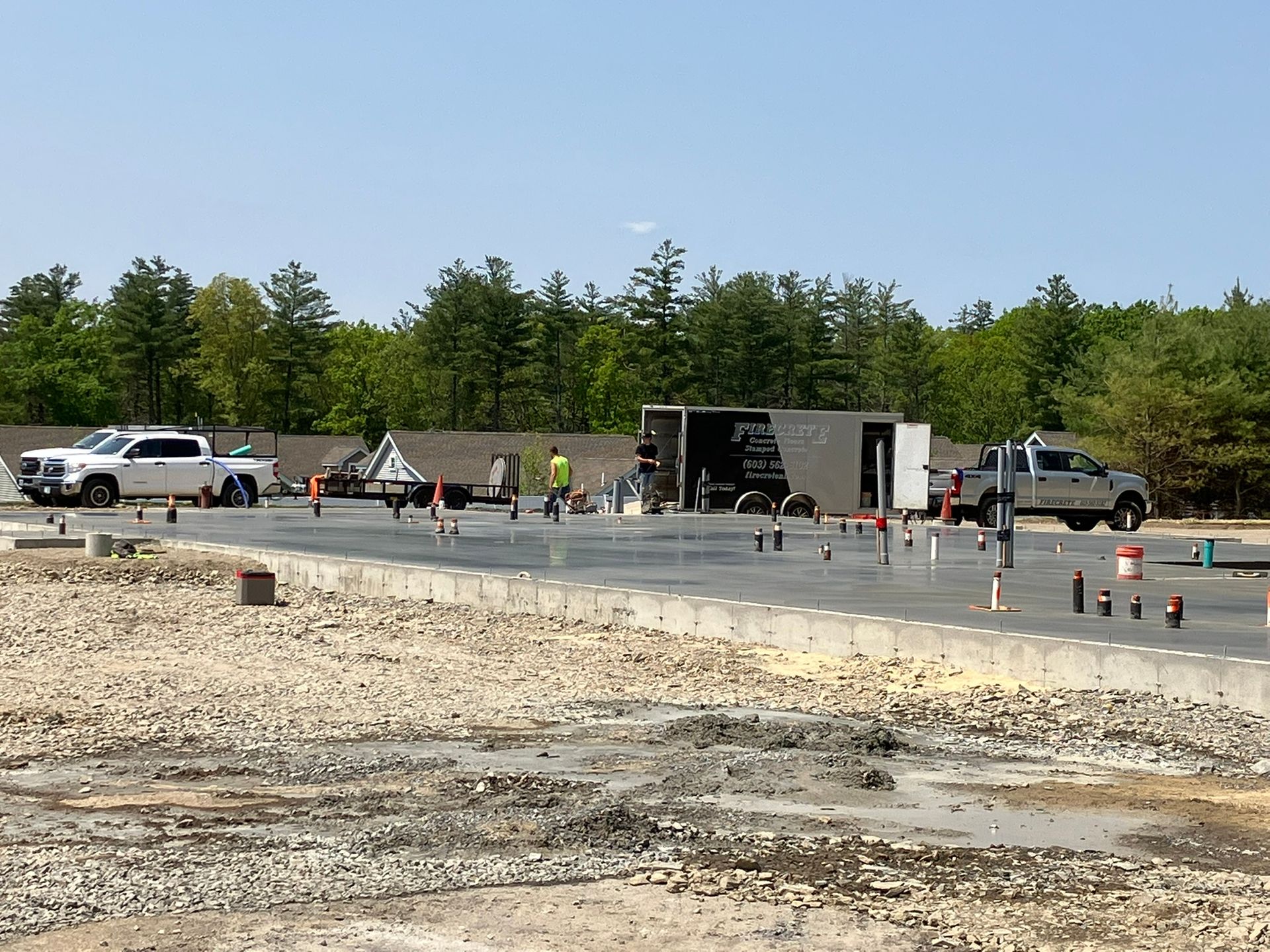 Construction workers pouring concrete; trucks, trees, and sky visible.
