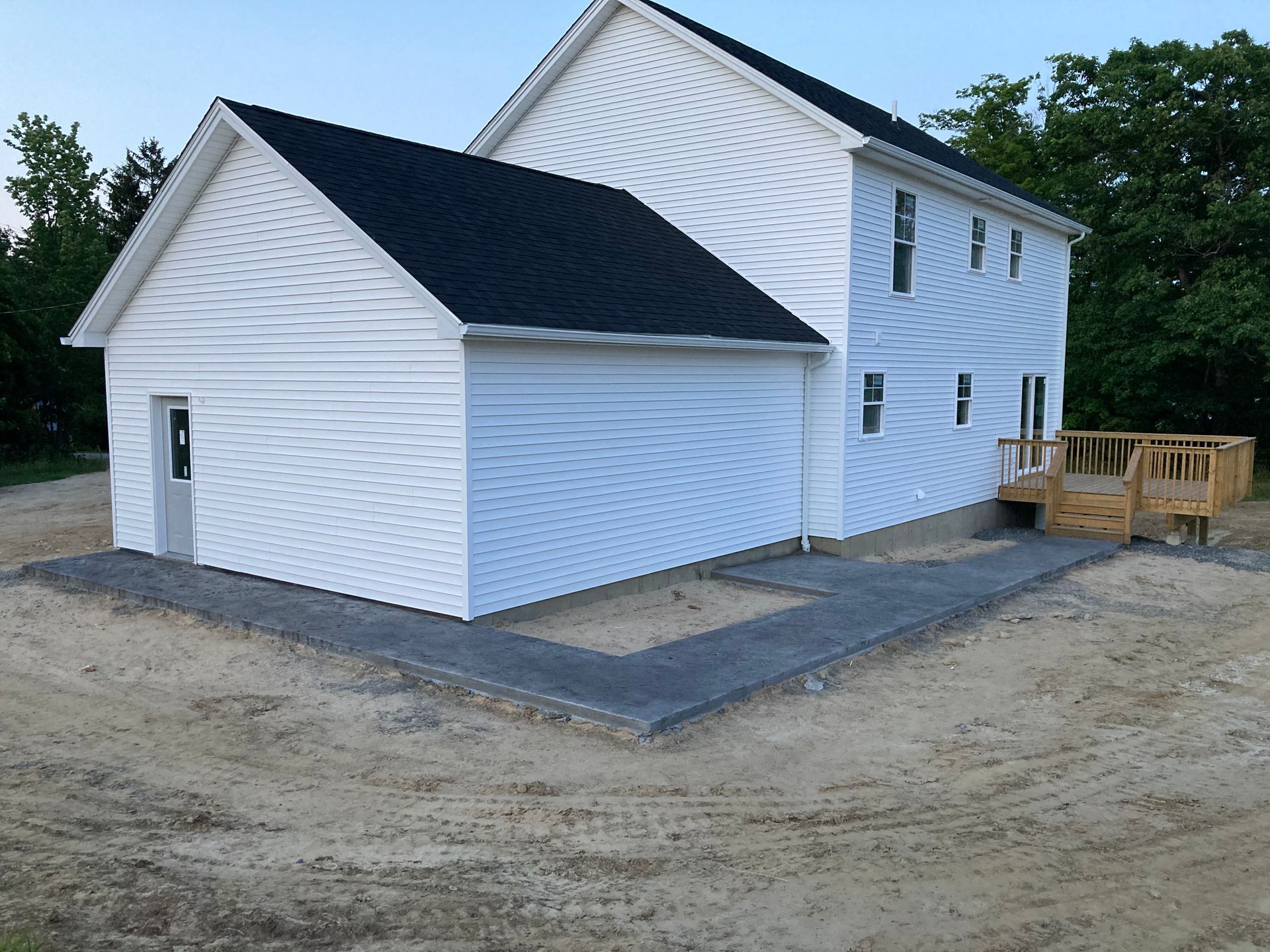 White house with attached garage, paved patio, and wooden deck; set on a dirt lot.