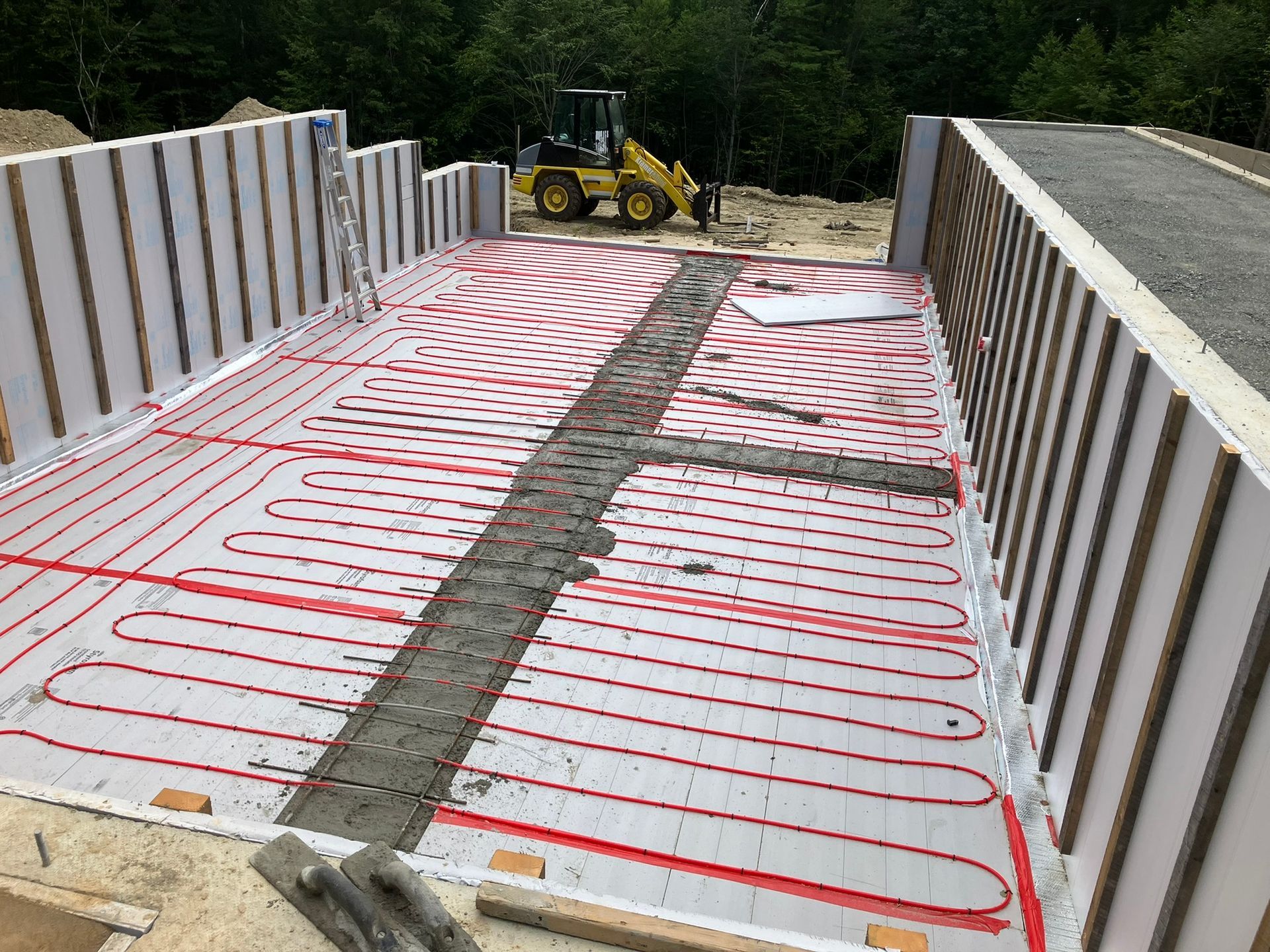 Construction site: radiant floor heating system with red tubing laid over white insulation, surrounded by concrete walls.