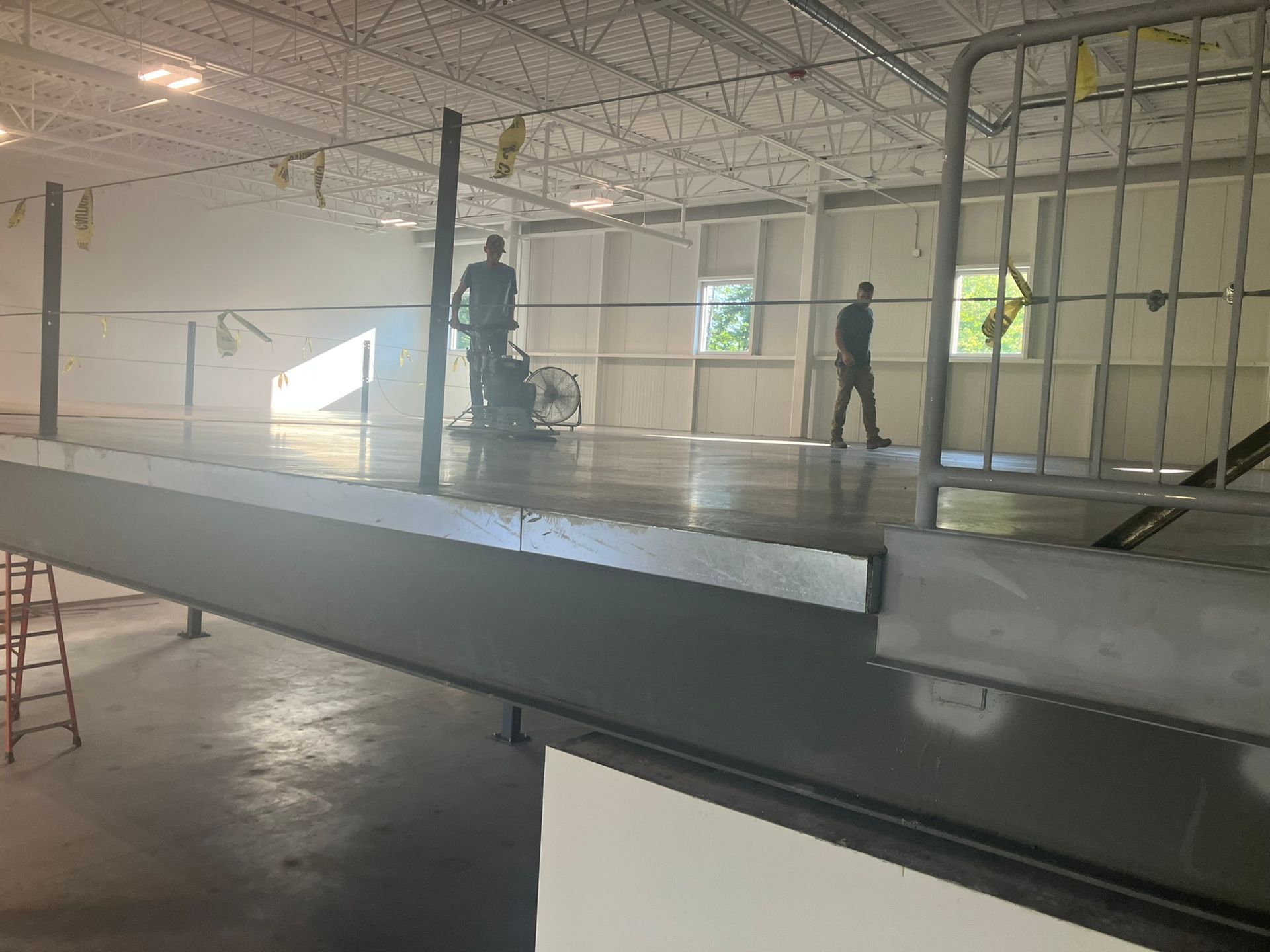 Two workers on a polished concrete floor; one operates a floor grinder, the other walks by. Large, open space.