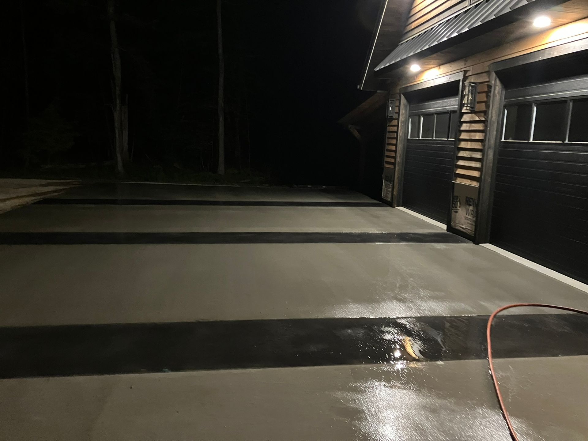 Nighttime view of a garage with black doors and concrete driveway with dark grey stripes; wet surface.