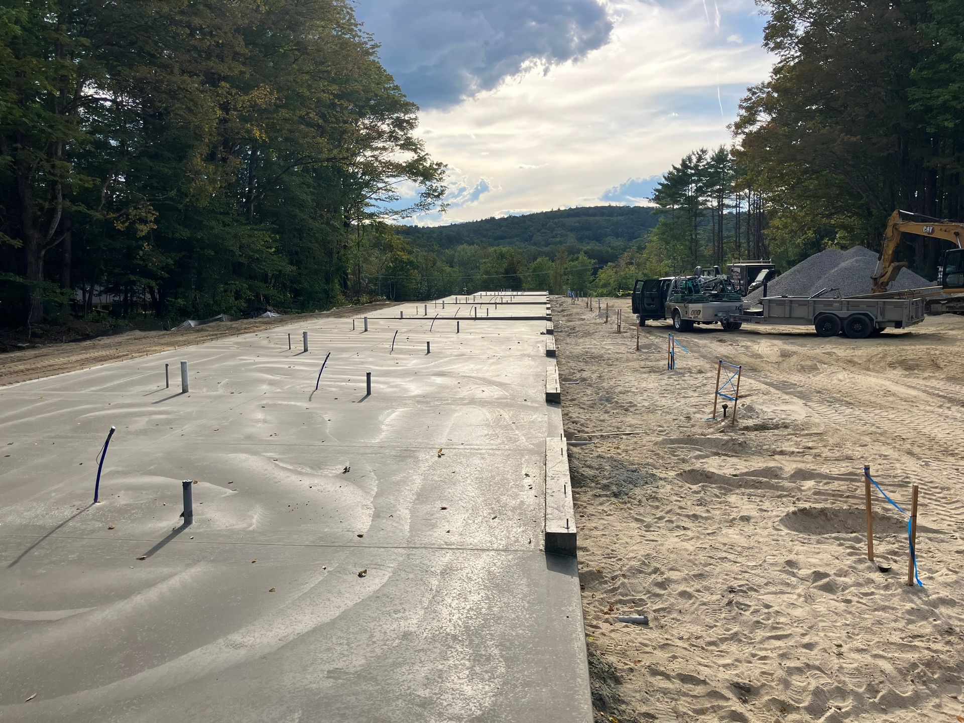 Newly poured concrete foundation with plumbing pipes, construction vehicles, and forest in the background.