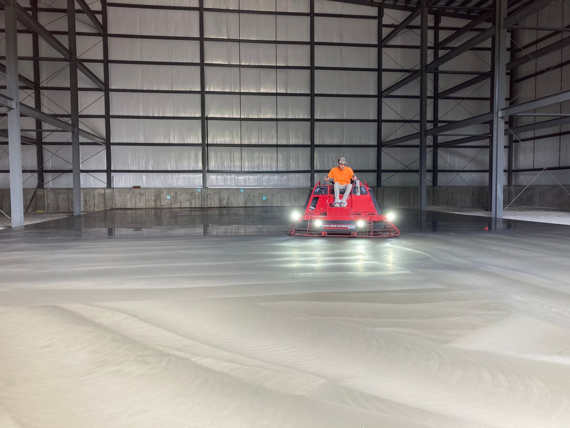 Man operating a red concrete finishing machine inside a large steel-framed building, smoothing a gray concrete floor.