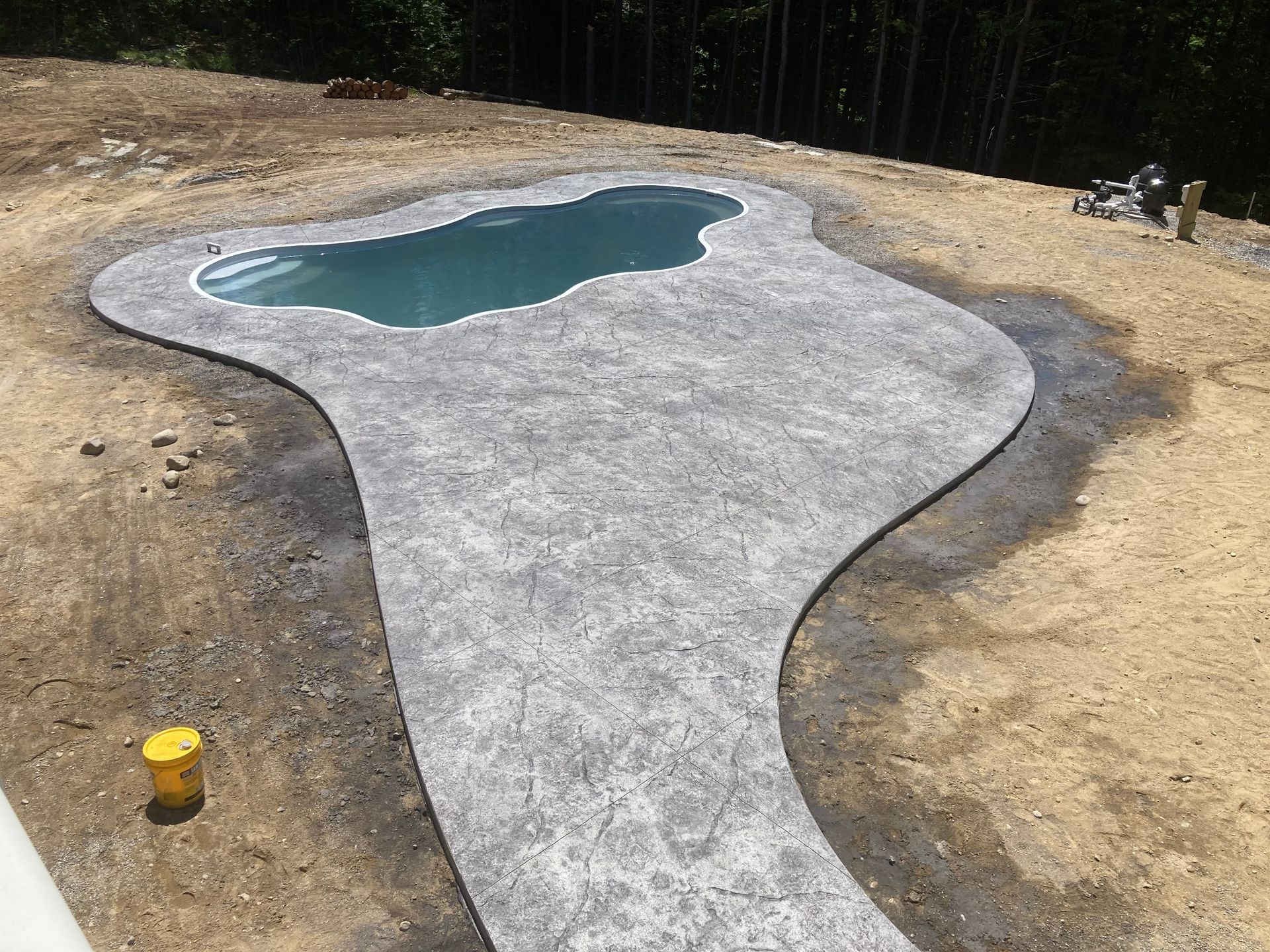A newly built, uniquely shaped pool with gray concrete surrounding it, set in a dirt yard.