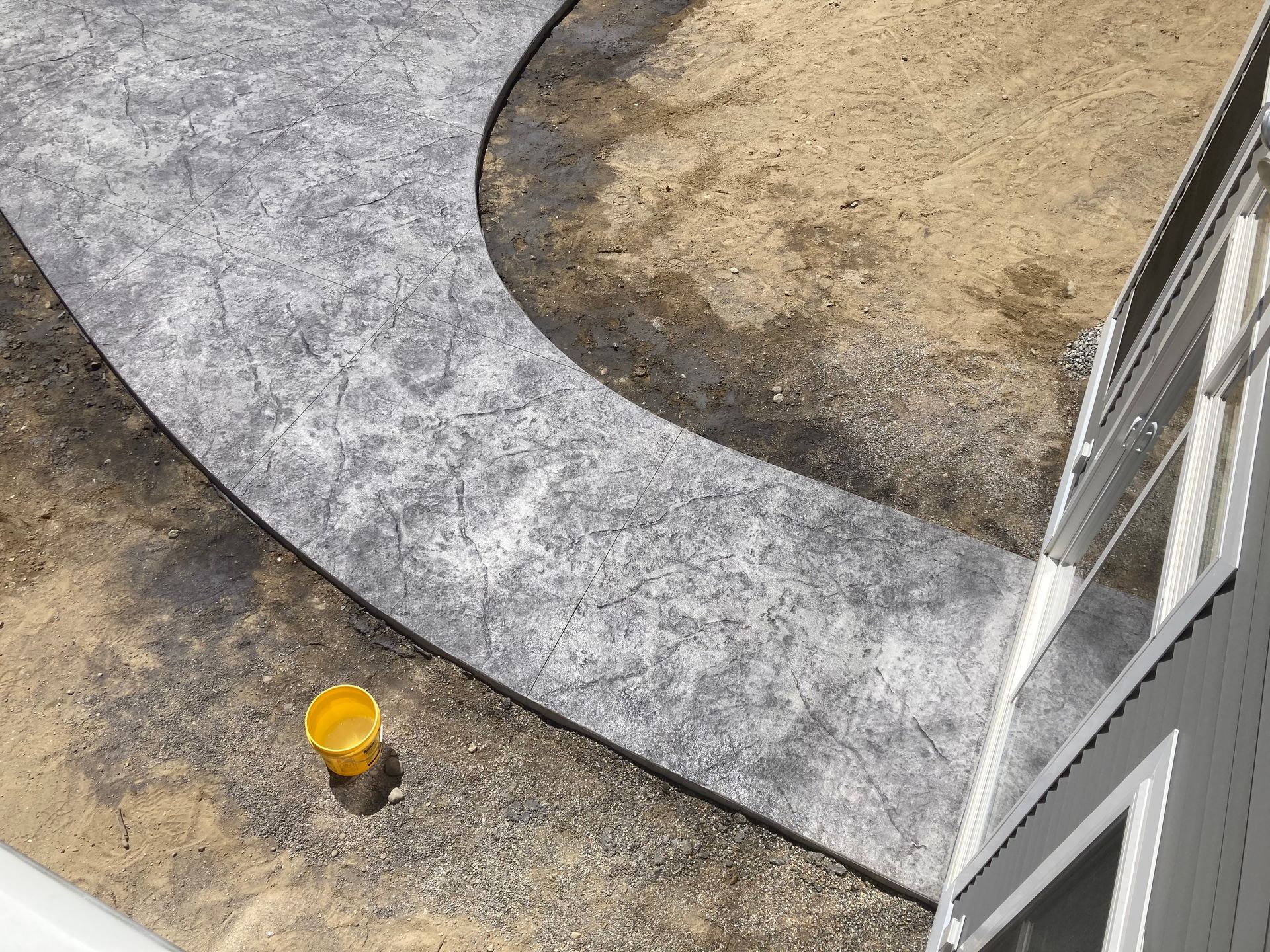 Gray textured concrete pathway curving through a dirt area next to a white house.