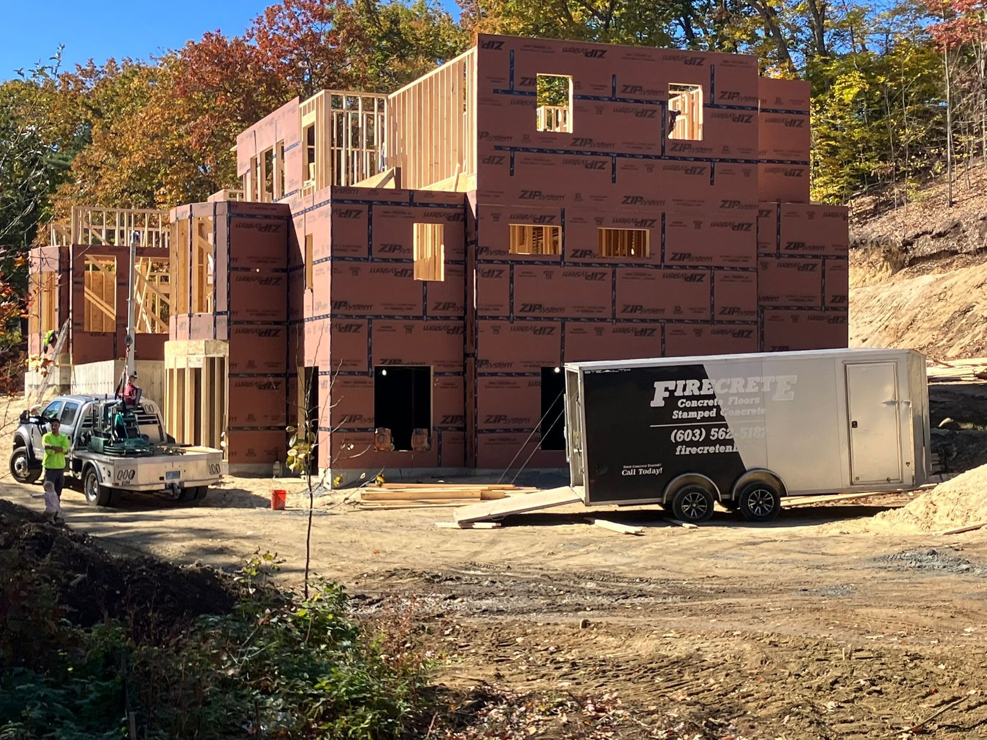 Construction site with house frame and siding, truck, trailer, and trees in the background on a sunny day.