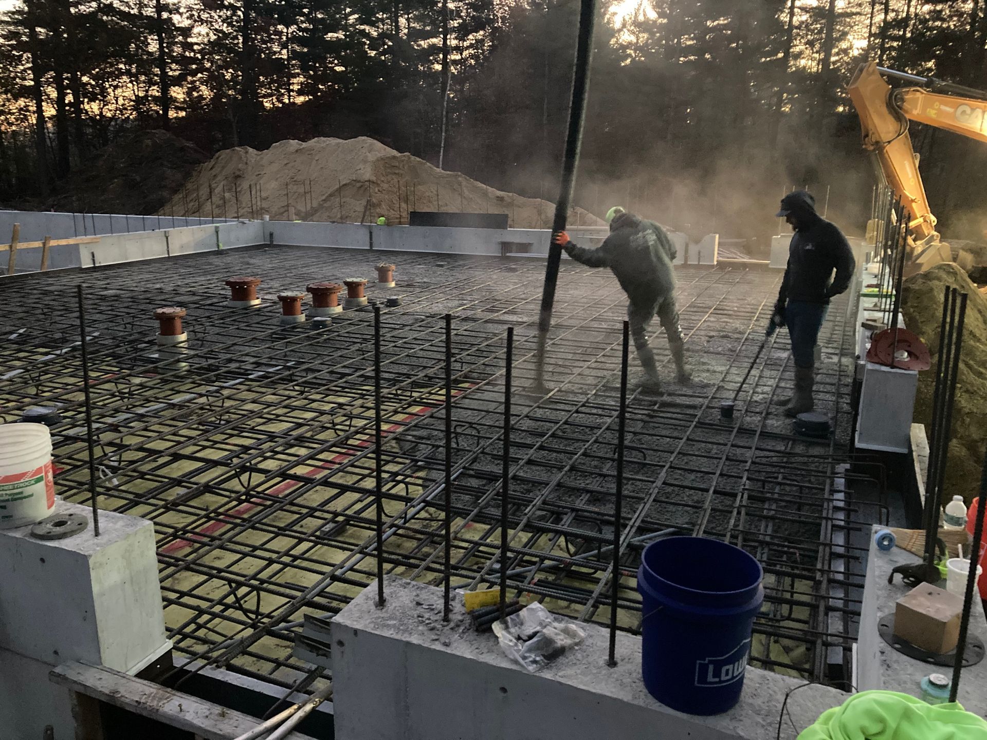 Workers pouring concrete foundation for a building. Dark sky and trees in background.