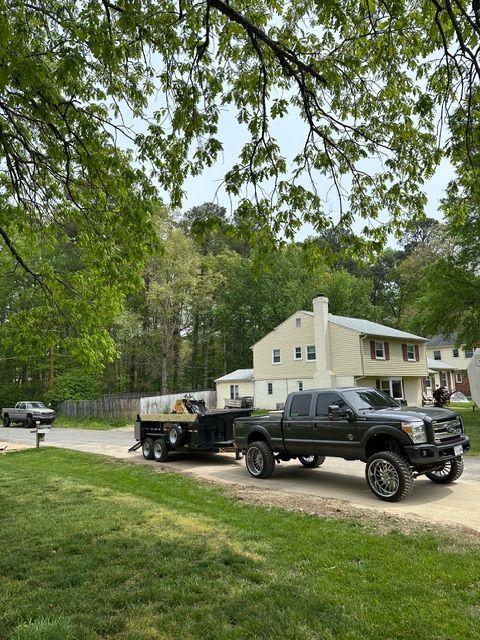 Black pickup truck towing a flatbed trailer on a gravel driveway beside a white house and trees