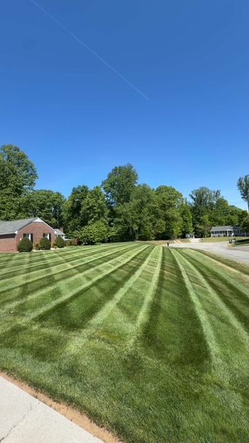 Freshly mowed grassy field with diagonal stripes beside a sidewalk, trees, and a small brick building under a blue sky