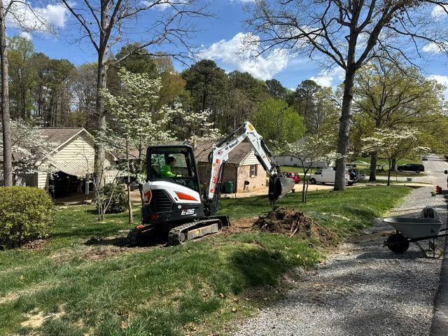 Mini excavator clearing a grassy roadside area near houses under a bright blue sky