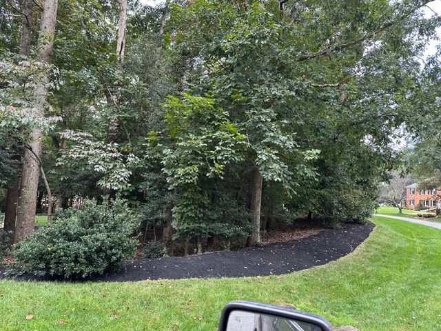 Tree-lined park path beside a dark mulch bed and green lawn, with houses in the background.