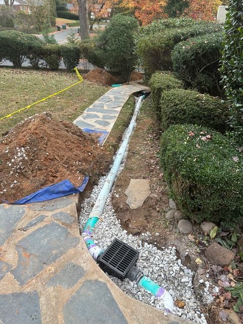 Broken stone walkway beside a drainage trench, with rubble, gravel, and shrubs in a landscaped yard.