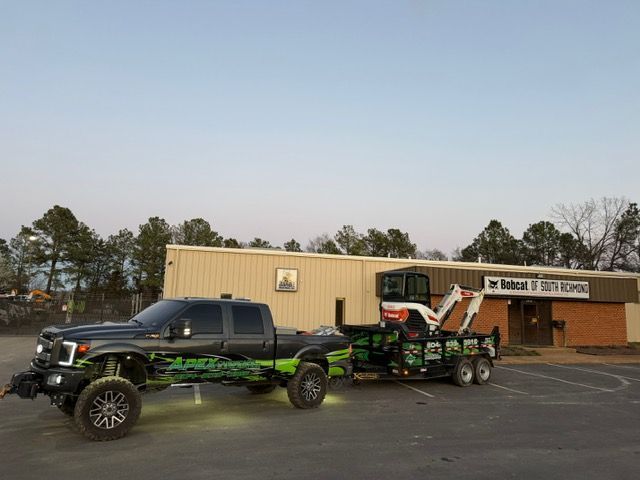 Black lifted pickup truck towing a custom trailer with orange off-road equipment in a parking lot outside a building