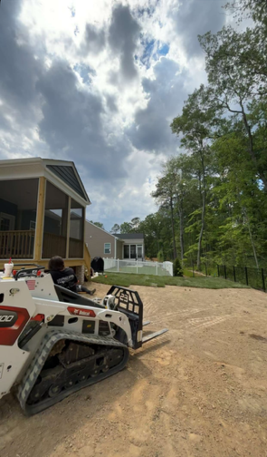 Person on a white skid steer beside a house on a dirt driveway under cloudy skies