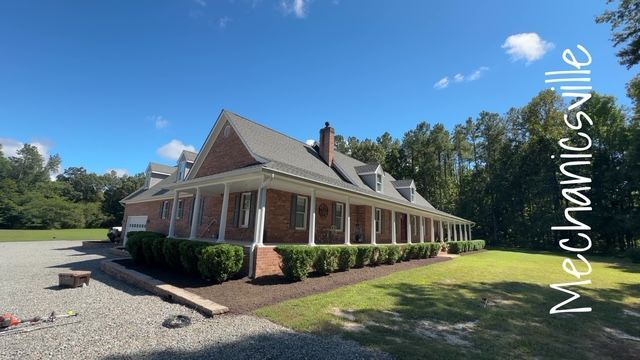 Brick house with a wraparound porch, white trim, and a gravel driveway on a sunny day
