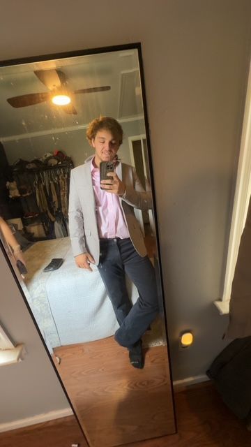 Person taking a mirror selfie in a bedroom, wearing a light blazer and pink shirt, with a warm lamp on the floor.