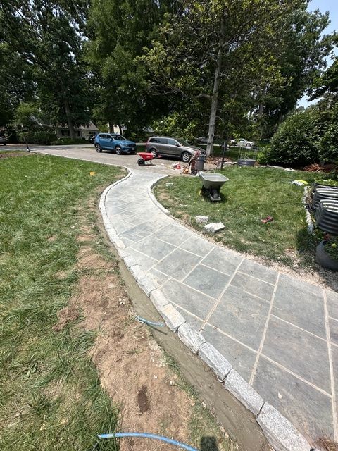 Curved stone walkway through a grassy yard with trees, parked cars, and a small red toy cart nearby