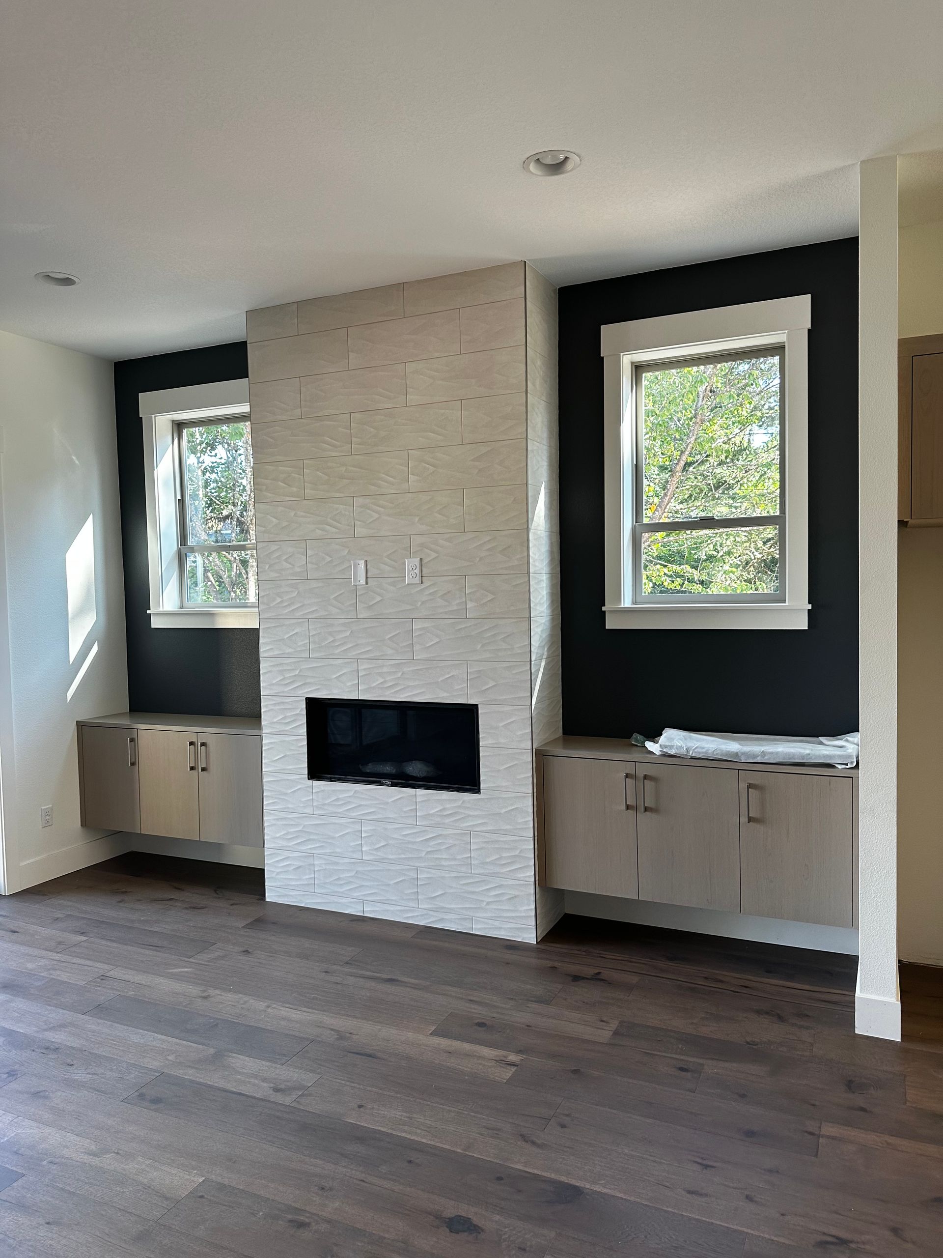Living room with a fireplace, built in cabinets, dark walls, and two windows.