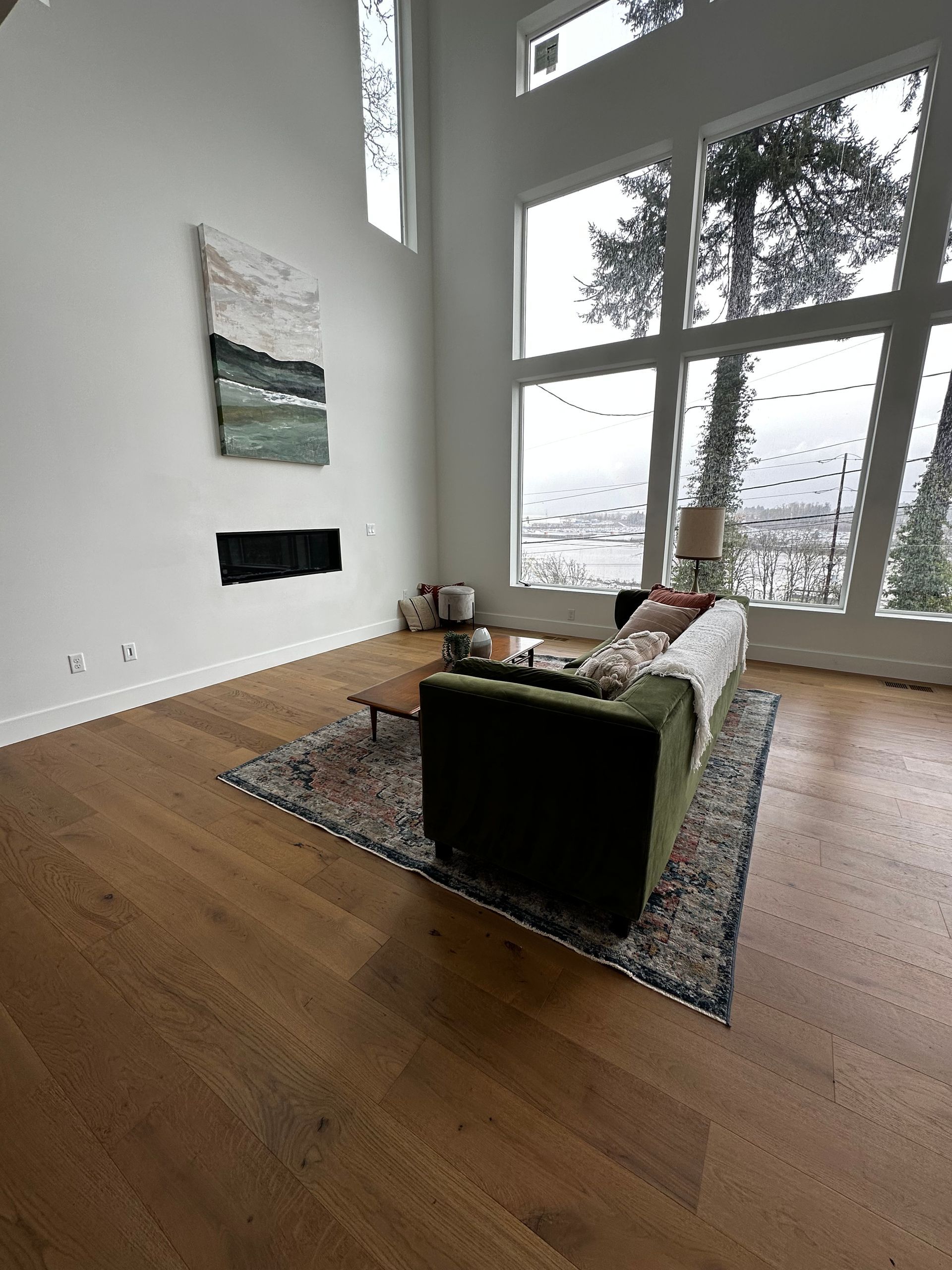Living room with tall windows overlooking trees and water. Green couch, rug, fireplace, and painting.