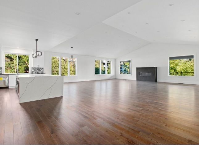 Spacious, empty living area with wooden floor, white walls, and large windows overlooking trees.
