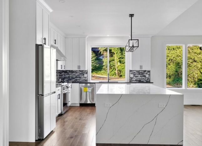 Modern white kitchen with island, stainless steel appliances, and forest view windows.