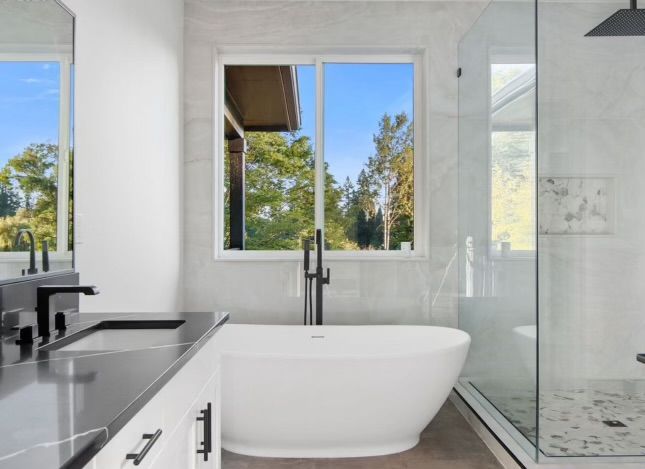 Modern bathroom with white tub, black fixtures, and large window overlooking trees.