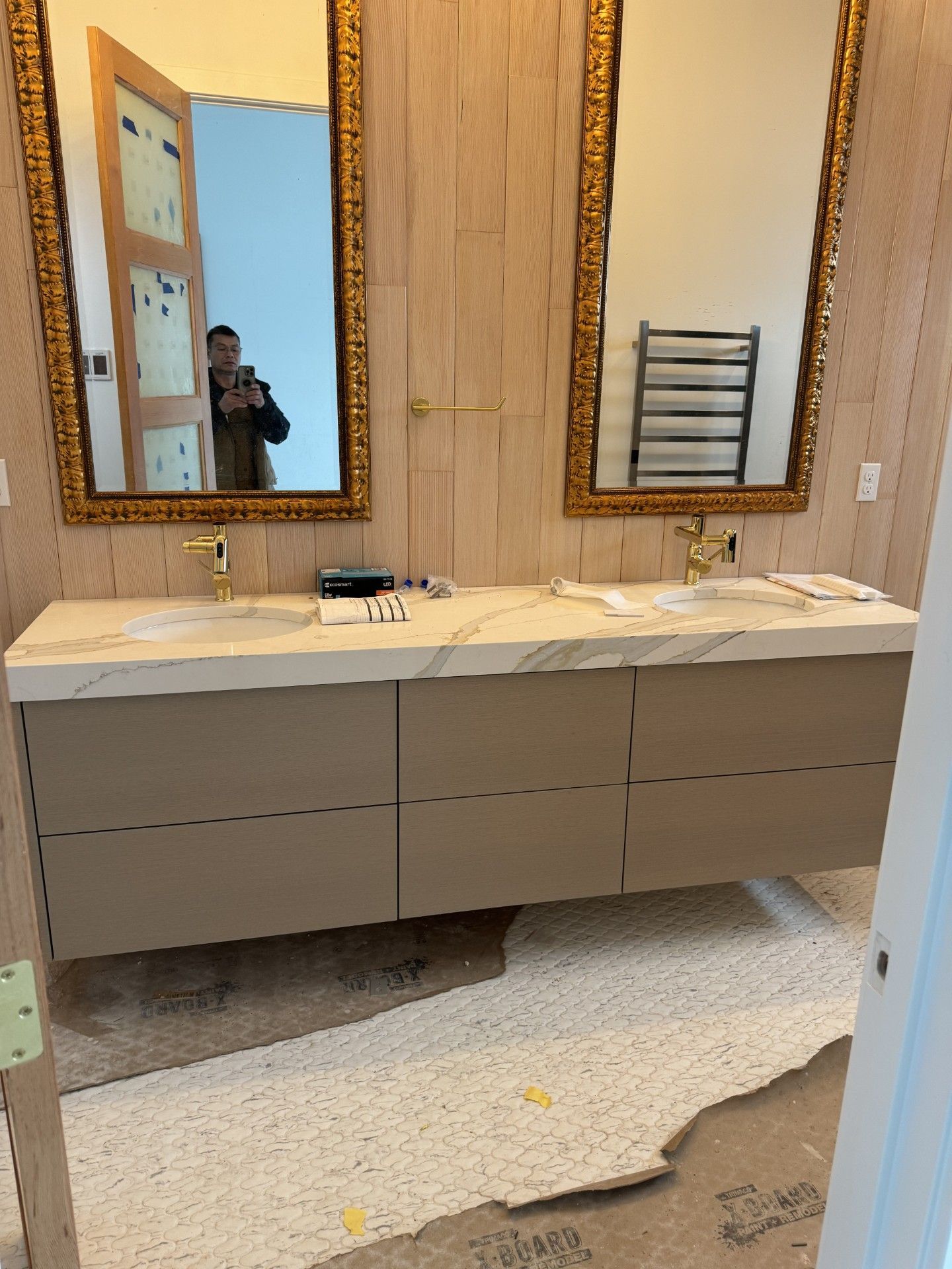 Bathroom with dual sinks, gold framed mirrors, beige vanity, and unfinished flooring.