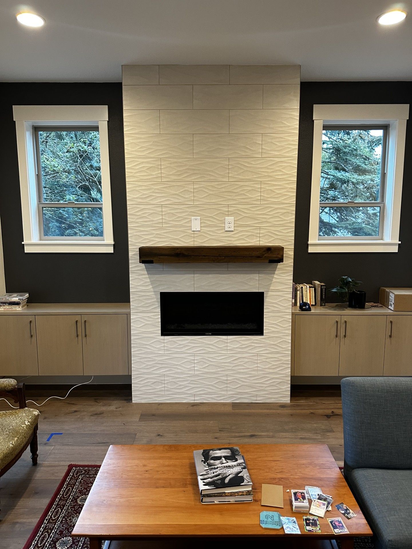 Living room with a white stone fireplace, dark cabinets, and windows with white trim.