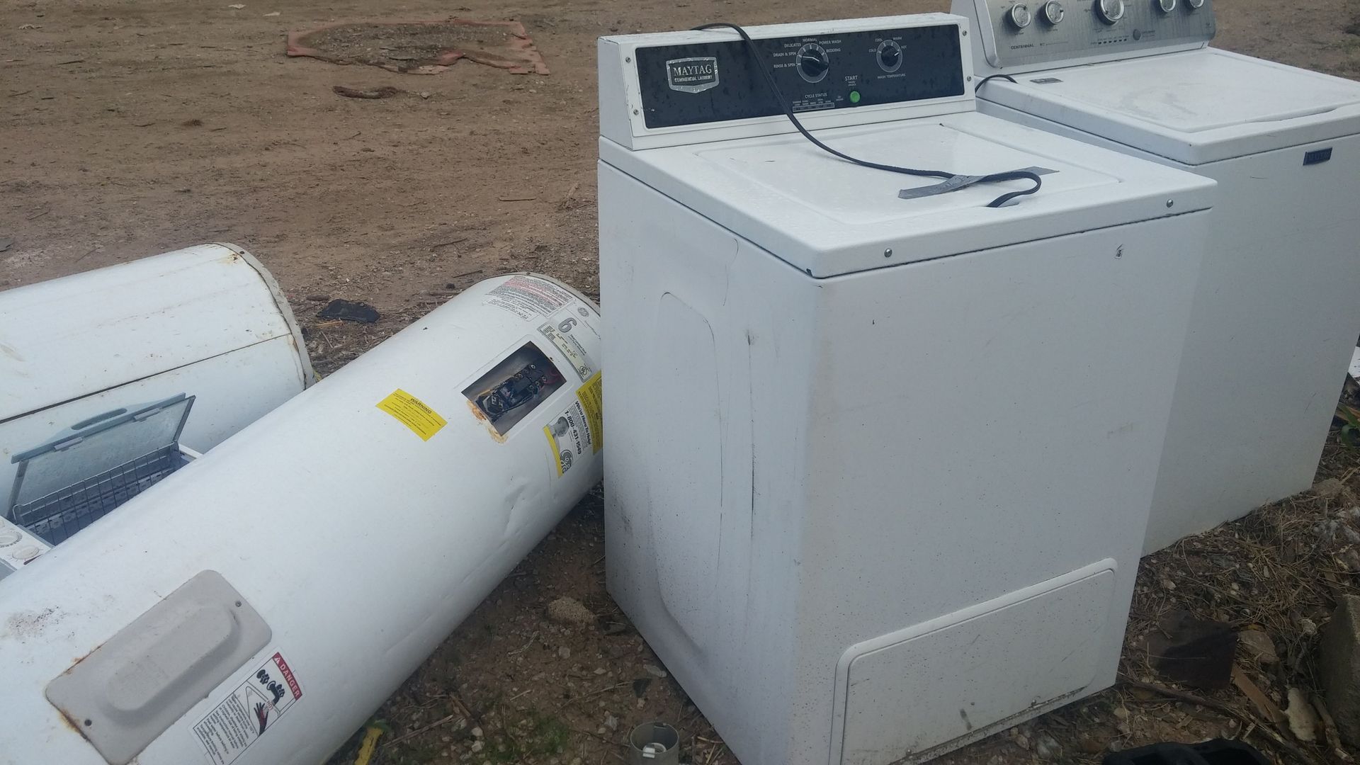 White dryer, washing machine, and water heaters on dirt ground