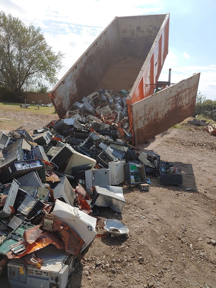 A large orange dumpster overflowing with discarded computer parts and electronic waste on a gravel surface