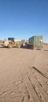 A yellow front-end loader lifts a trailer with a large green container across a sandy field under a blue sky