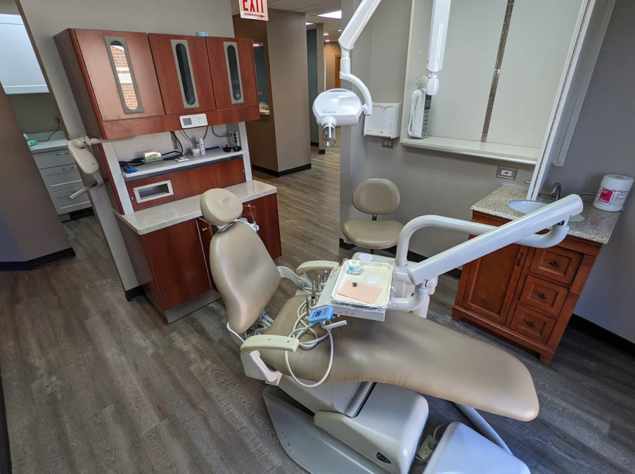 Dental clinic operatory with beige exam chair, overhead light, sink, and wooden cabinetry.