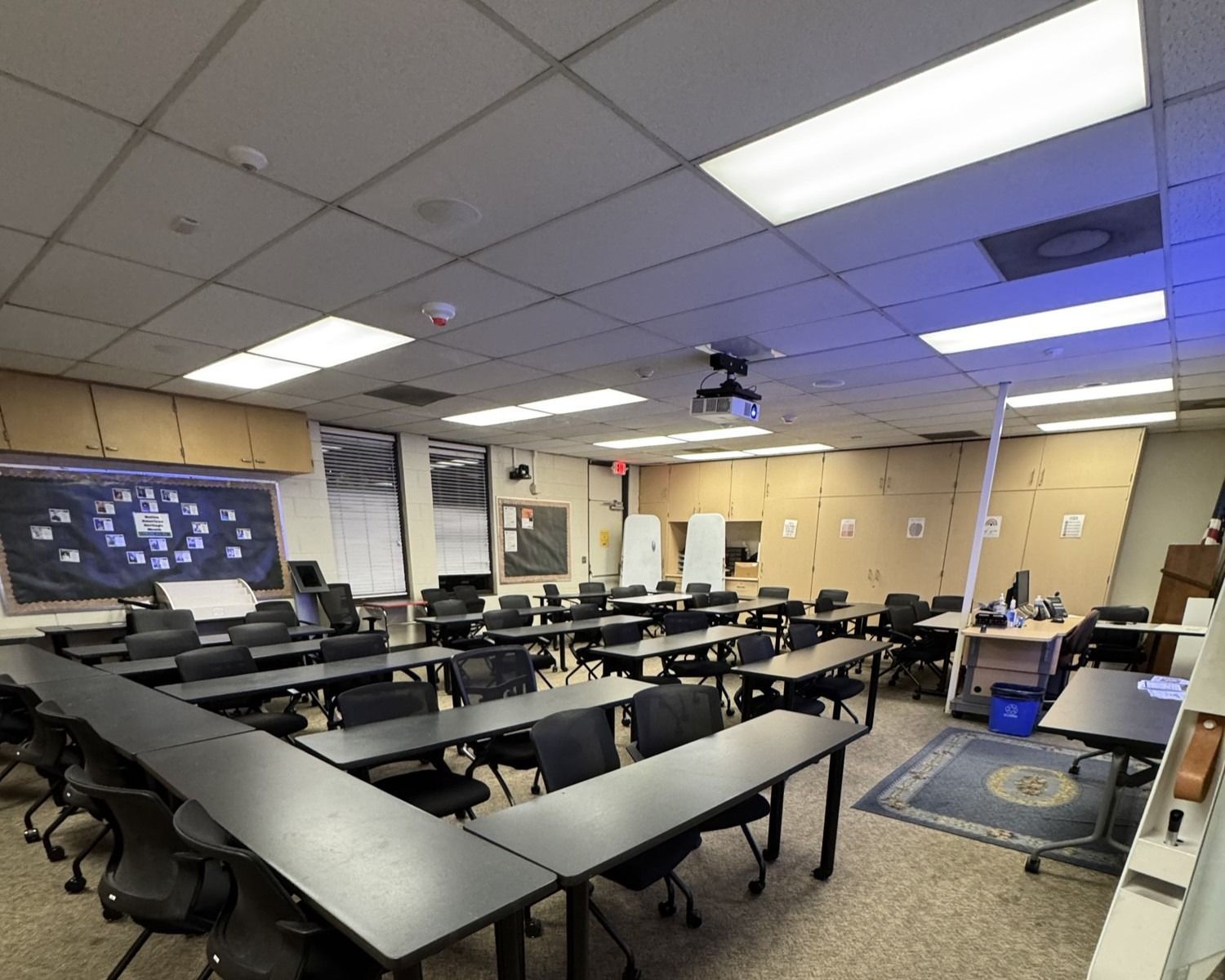 Empty classroom with U-shaped black tables, chairs, ceiling lights, and a whiteboard at the front