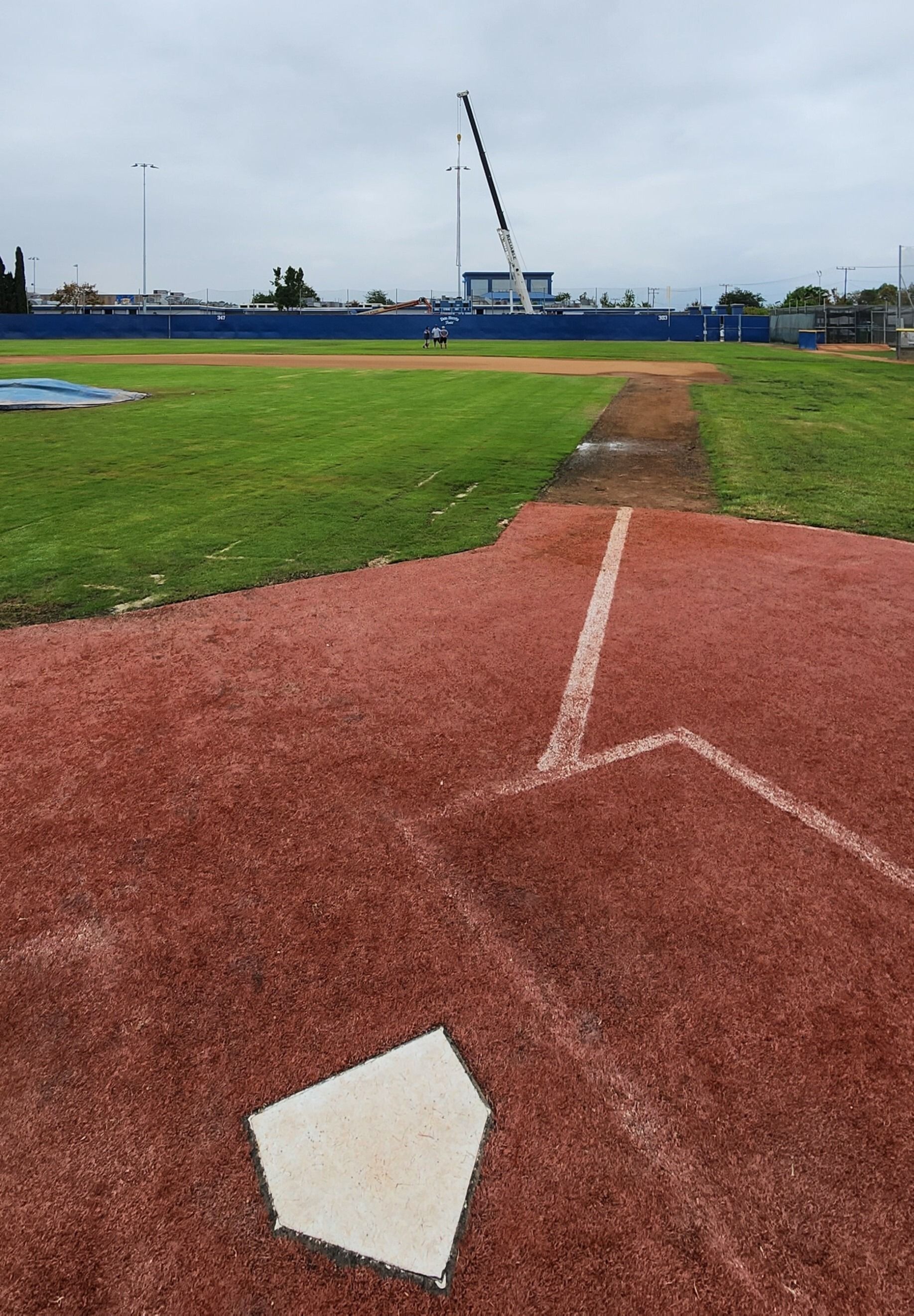 Baseball field view from home plate toward the outfield, with dirt baseline and green grass under cloudy sky.