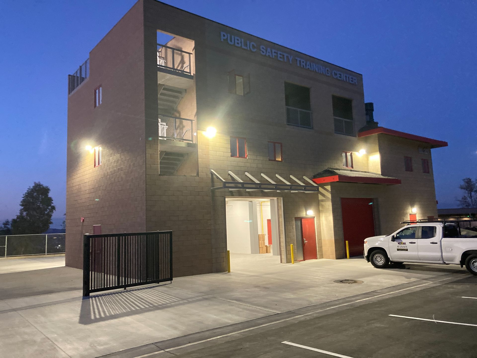 Modern two-story building at dusk with lit entrance, parked white pickup, and black fence in front