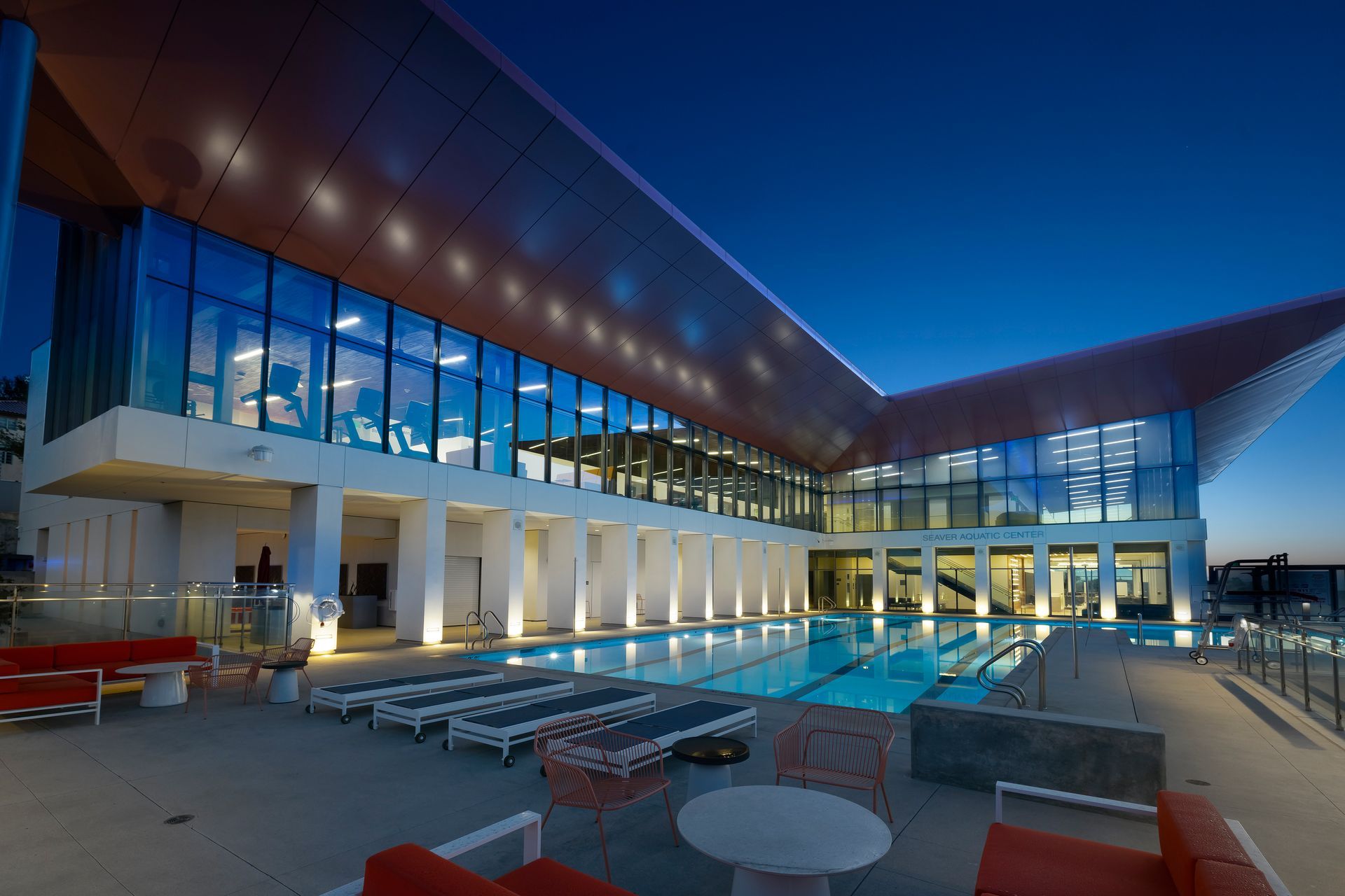Modern building courtyard at dusk with glass walls, a reflecting pool, and outdoor seating