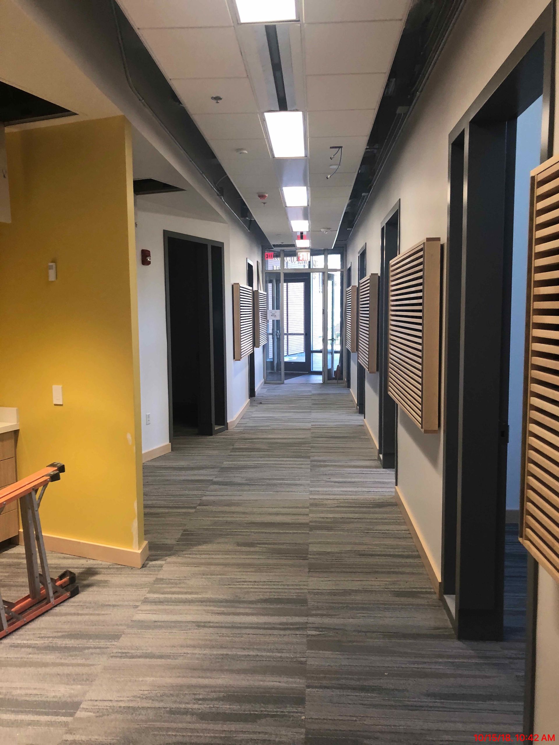 Empty office hallway with gray carpet, black doorways, yellow wall accent, and slatted wood panels.