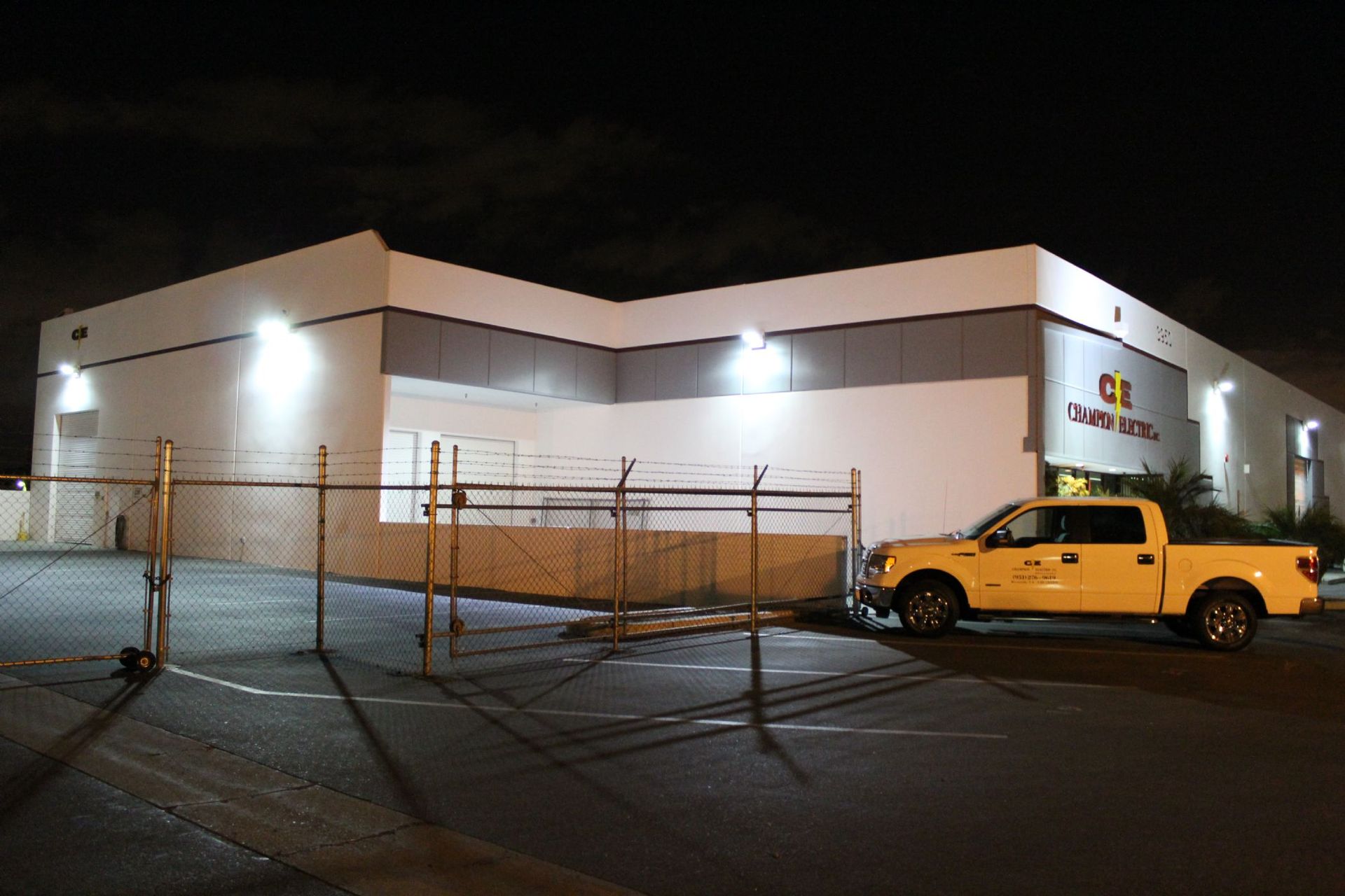 Night view of a white building with a fenced lot and a yellow pickup truck parked outside