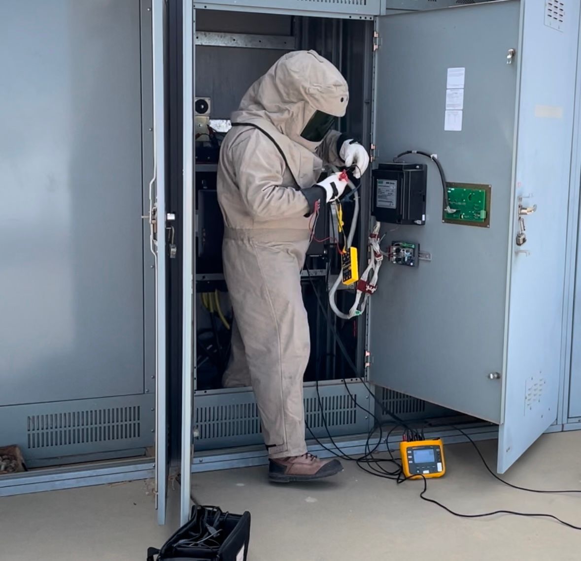 Person in protective coveralls inspecting electrical equipment inside an open cabinet.