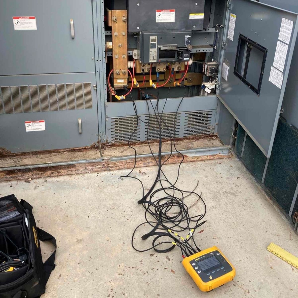 Open electrical panel with wires and a yellow meter on the floor in a utility room