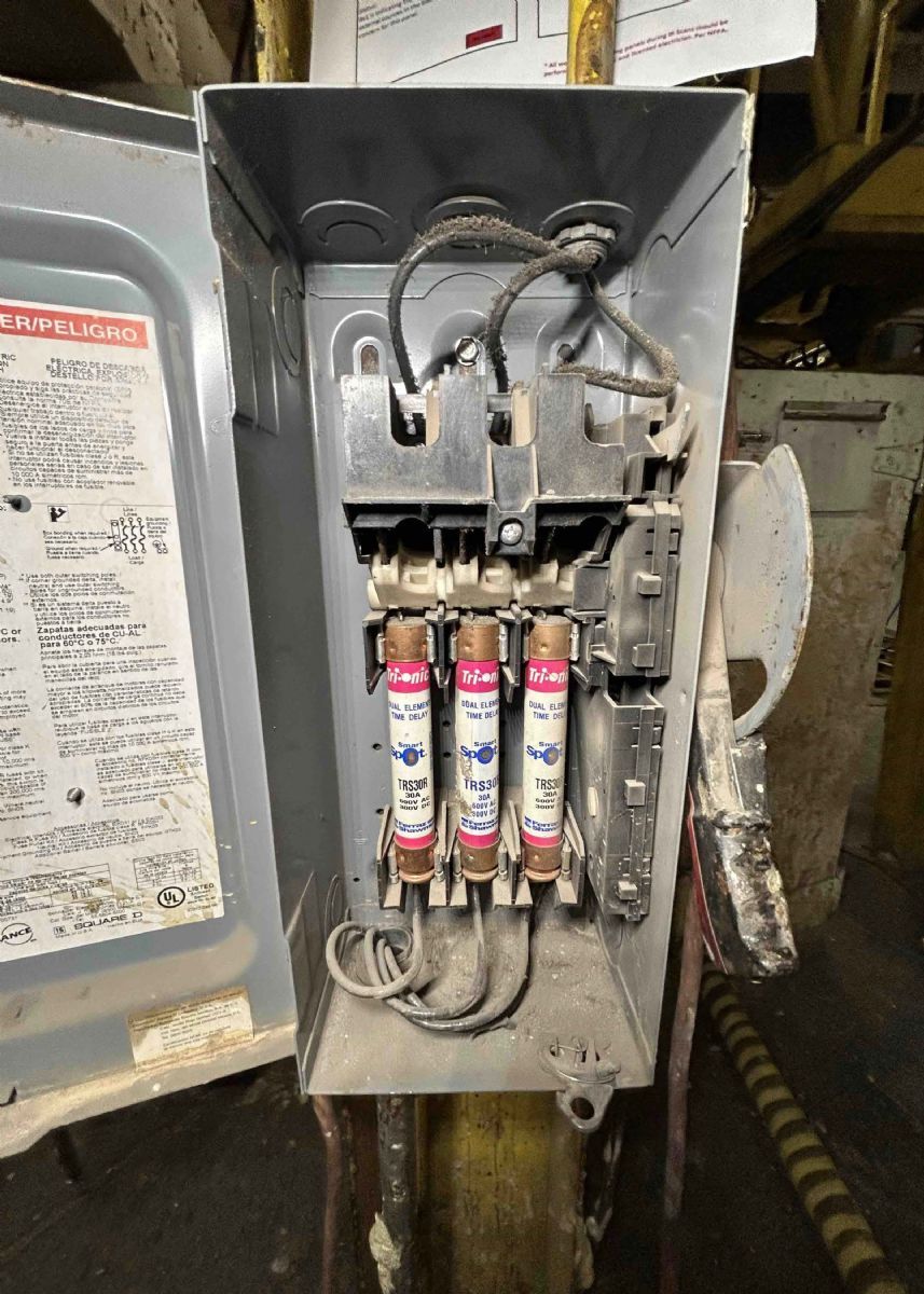 Open electrical fuse panel with three cylindrical fuses and exposed wiring inside a gray metal box.