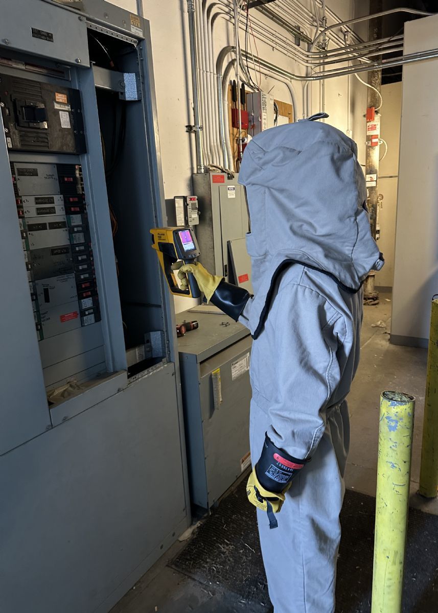 Person in blue hazmat suit inspecting electrical panels in a utility room.