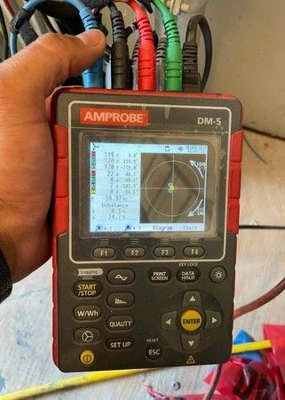 Hand holding an Amprobe DM-5 meter with cables connected and a display showing a circular graph.